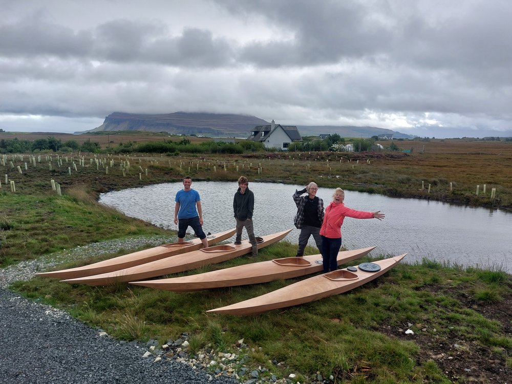 Four people standing by their newly built wooden kayaks, in a rugged Scottish island landscape