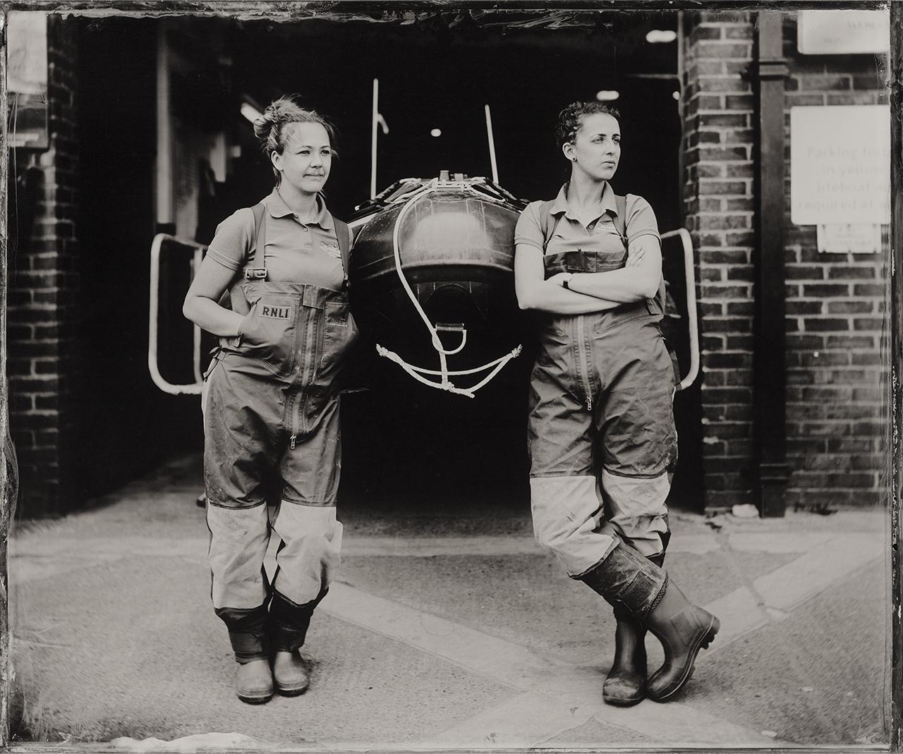 Black and white photo of two female RNLI volunteers in overalls, standing next to a lifeboat