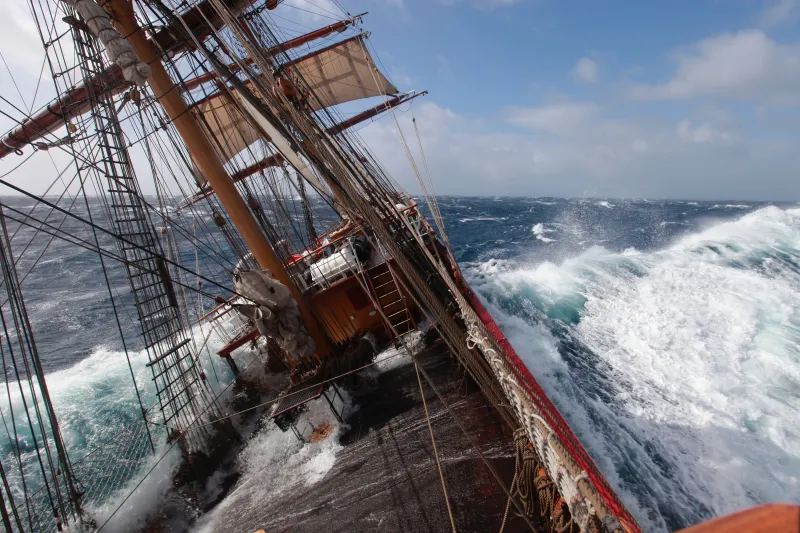 A view from over the gunwale of a tall ship looking along the ship's deck. The vessel is heeling to the left of the image at a steep angle, with deep blue waves foaming alongside it. The horizon ine in the distance is even, and the sky above light blue with some white clouds in the distance.