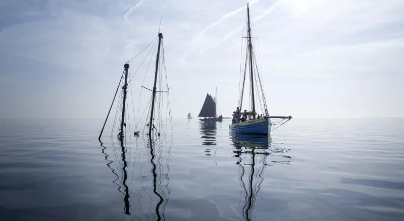 The sky melds with the grey water. They cover almost the whole image. In the centre, a starboard bow view of smack CK363 as she approaches a wreck with two masts sticking out of the water. In the background two more smacks can be seen, on their starboard sides.