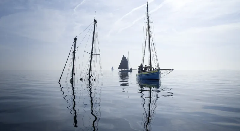 The sky melds with the grey water. They cover almost the whole image. In the centre, a starboard bow view of smack CK363 as she approaches a wreck with two masts sticking out of the water. In the background two more smacks can be seen, on their starboard sides.