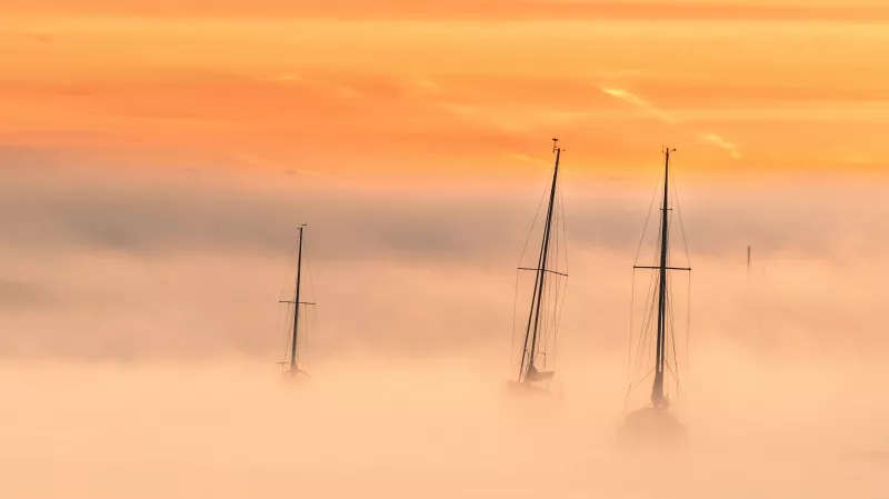 Orange hues cover the whole image. Bottom half of the vista is covered in fog, with three dark masts with stowed sails emerging from the fog in the centre of the photo.