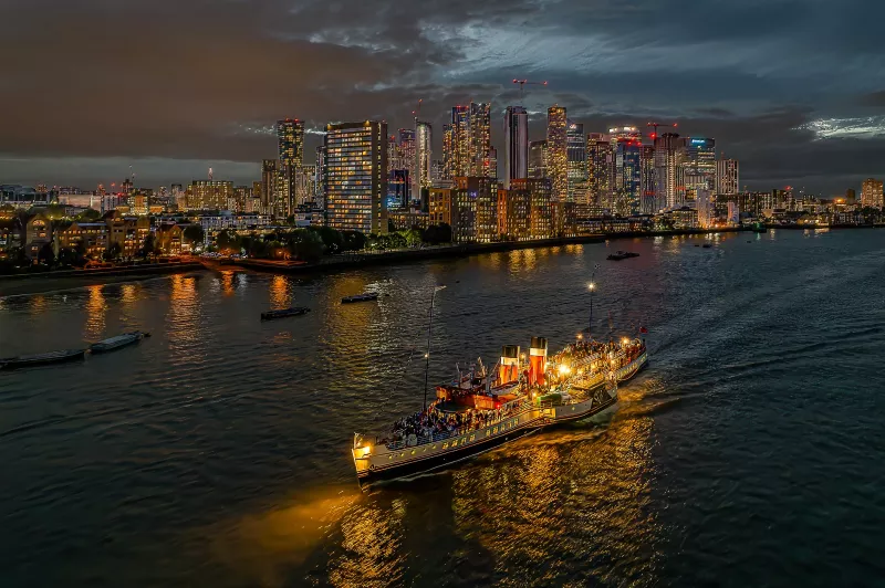 A birds eye view of the PS Waverley on the Thames after sundown, with the tall buildings of Canary Wharf in the background.