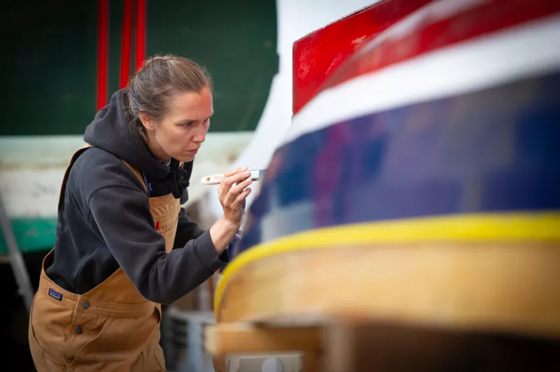 On the right part of an upturned hull of a boat is visible. It has colourful horizontal stripes. A woman on the left of the image is holding a brush to the hull, painting the boat.