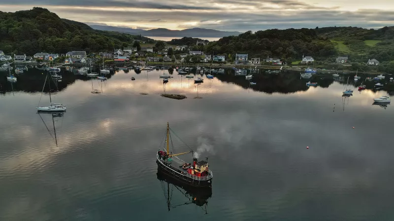 VIC 32 is shown in the centre foreground from port stern, approaching shore where multiple small yachts are moored. There are houses and dark trees beyond the water, and in the far distance mountain peaks can be seen.