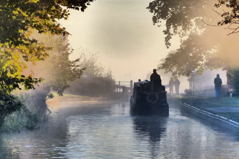Narrow boat sailing down a canal, trees overhanging the sides and a fog visible in the air