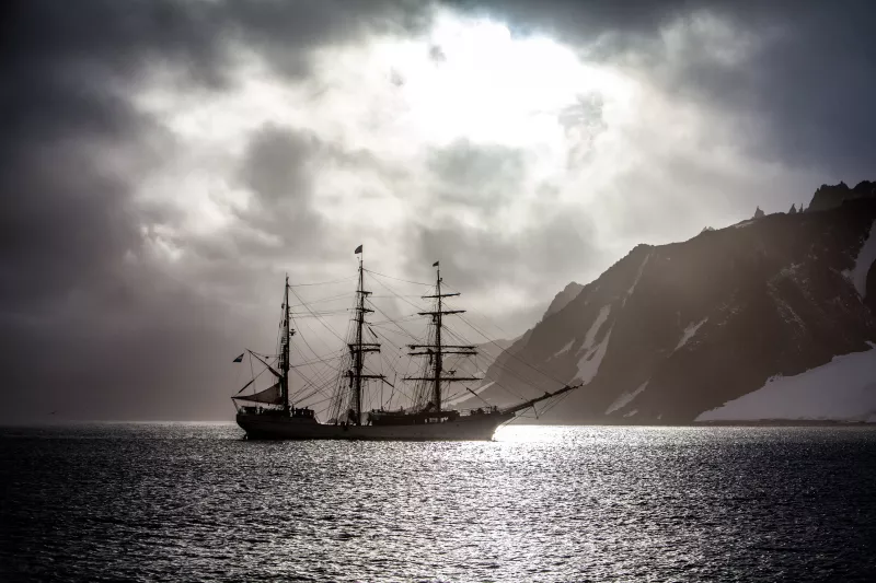 Bark Europa with all sails stowed against the backdrop of a snowy rock outcrop in Antarctica