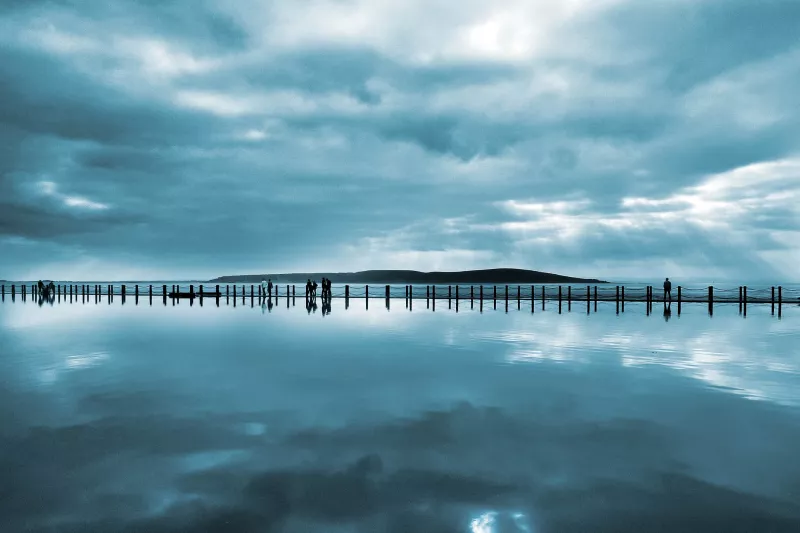 Distant view of a pier on a moody, cloudy day. The gloomy sky is reflected in the water.