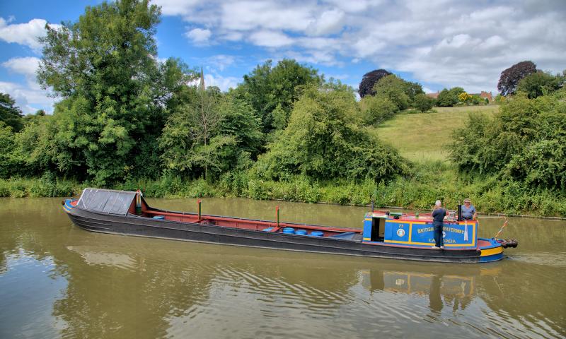 Photo Competition 2023 - Narrow boat Cassiopeia cruising along the Grand Union Canal with the spire of Braunston's St. Bartholomew's Church as a backdrop by Kev Maslin